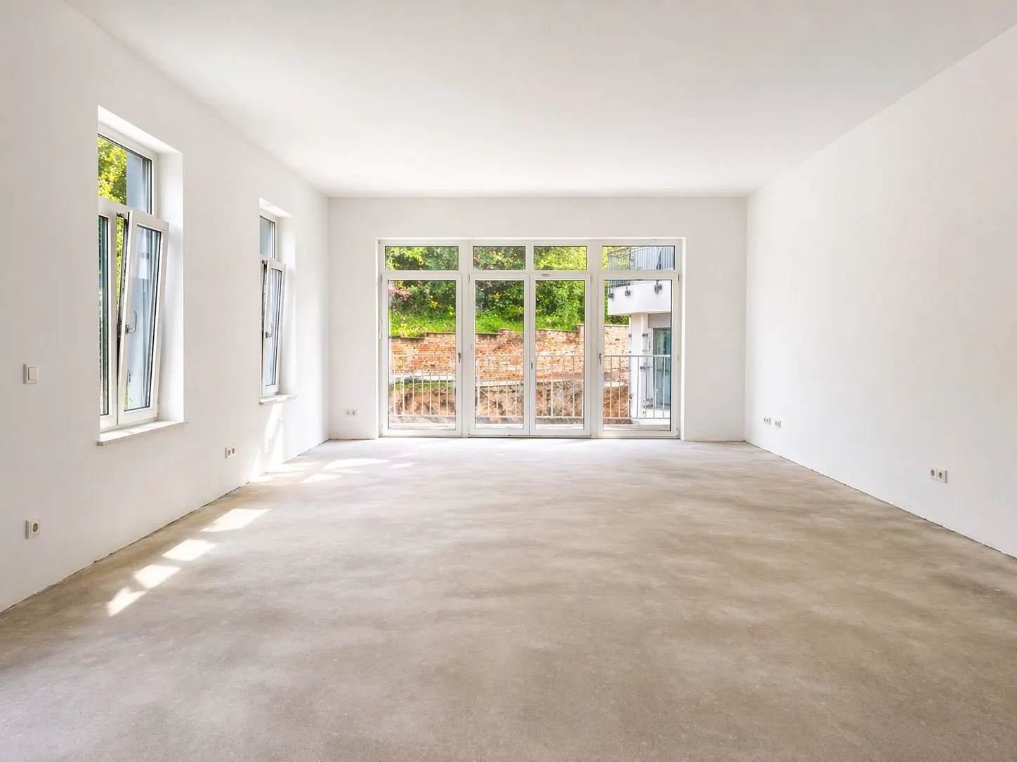 Heller, leerer Raum mit großen Fenstern und Blick auf eine Terrasse mit Garten.