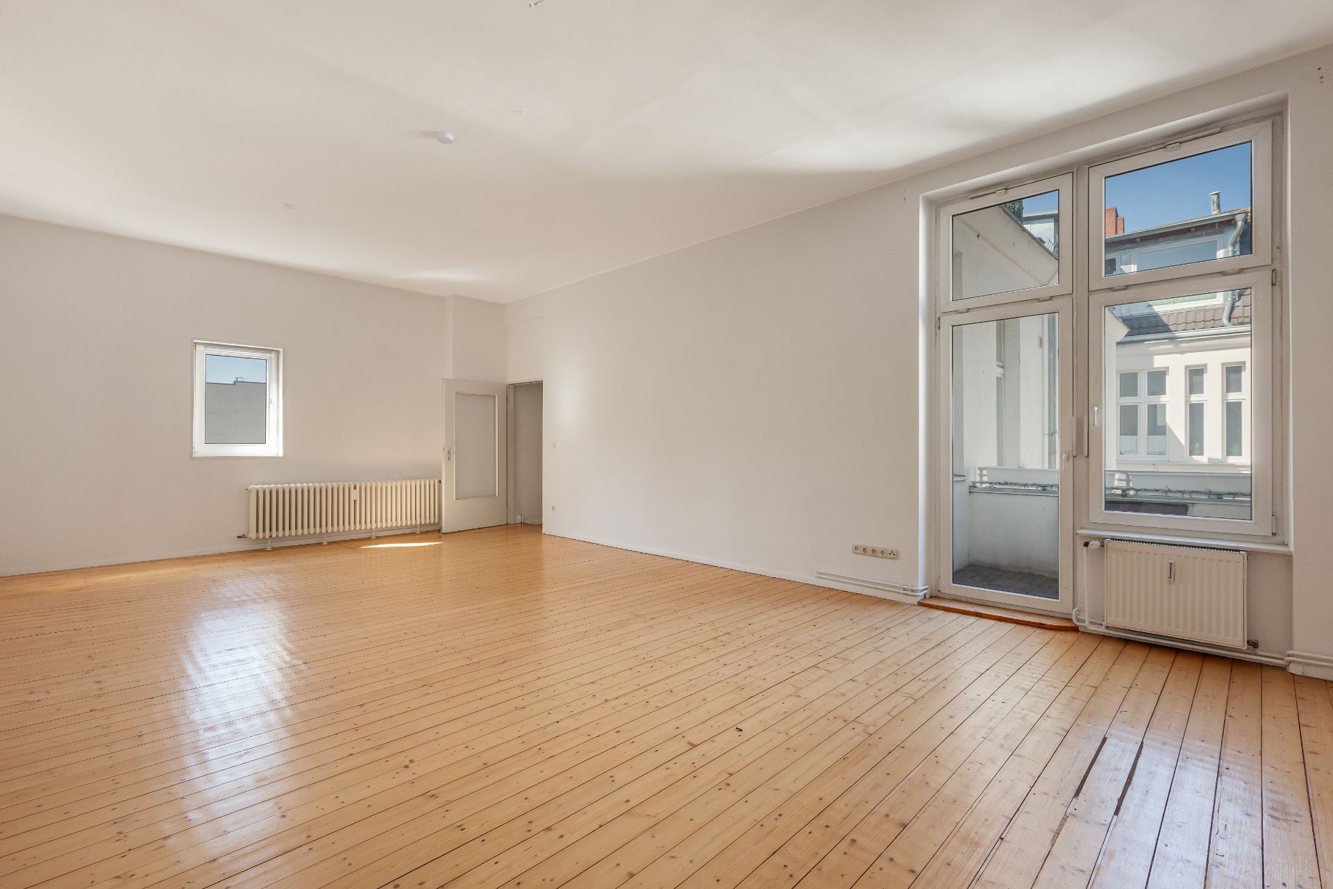 Helles, leeres Wohnzimmer mit Holzboden und großen Fenstern, Blick auf benachbarte Gebäude.