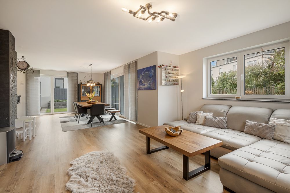 Helles Wohnzimmer mit Ecksofa, Holztisch und Essbereich, große Fenster und Holzboden.