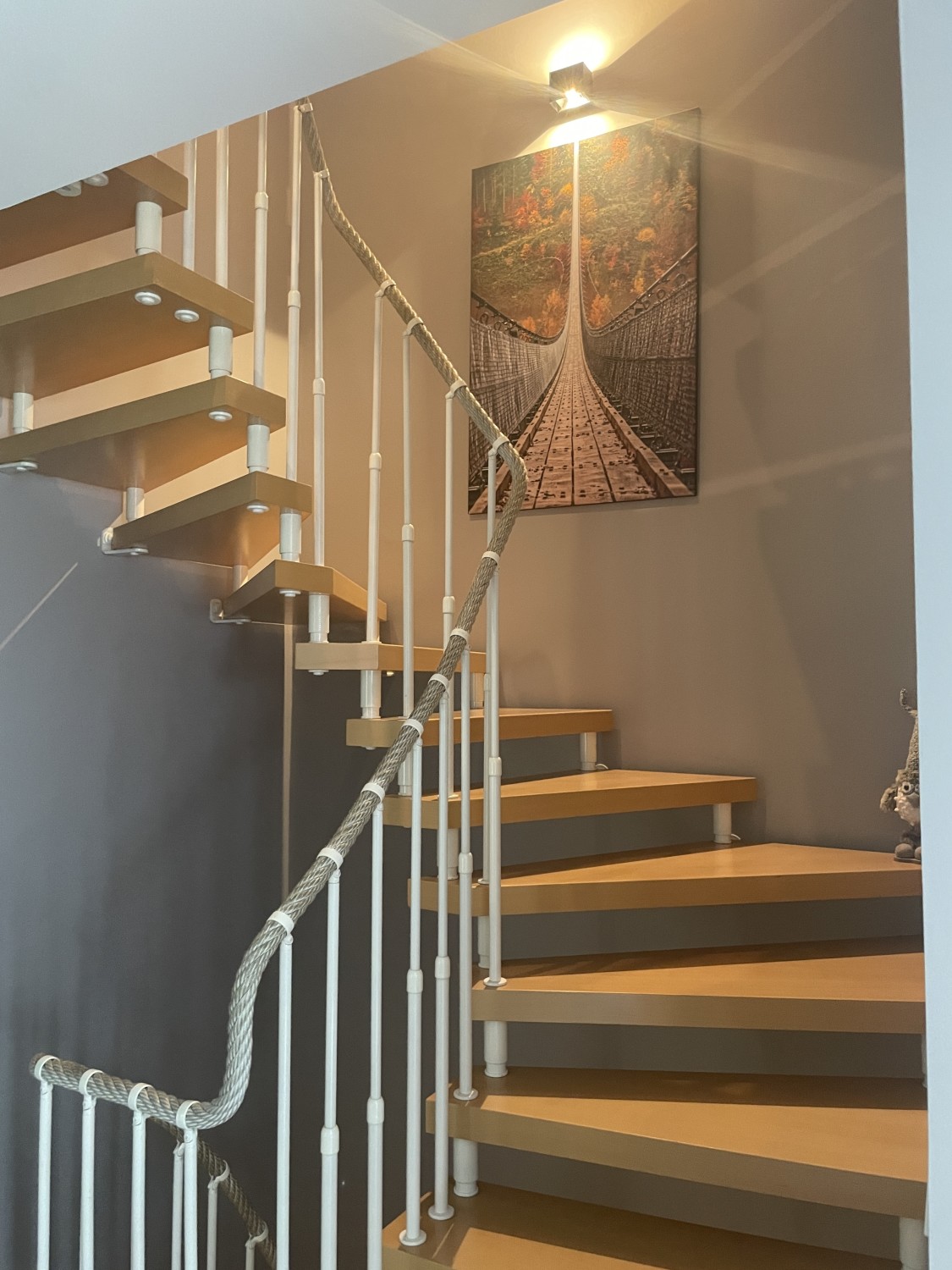 Innenansicht einer Holztreppe mit weißem Geländer und Wandbild einer Hängebrücke.