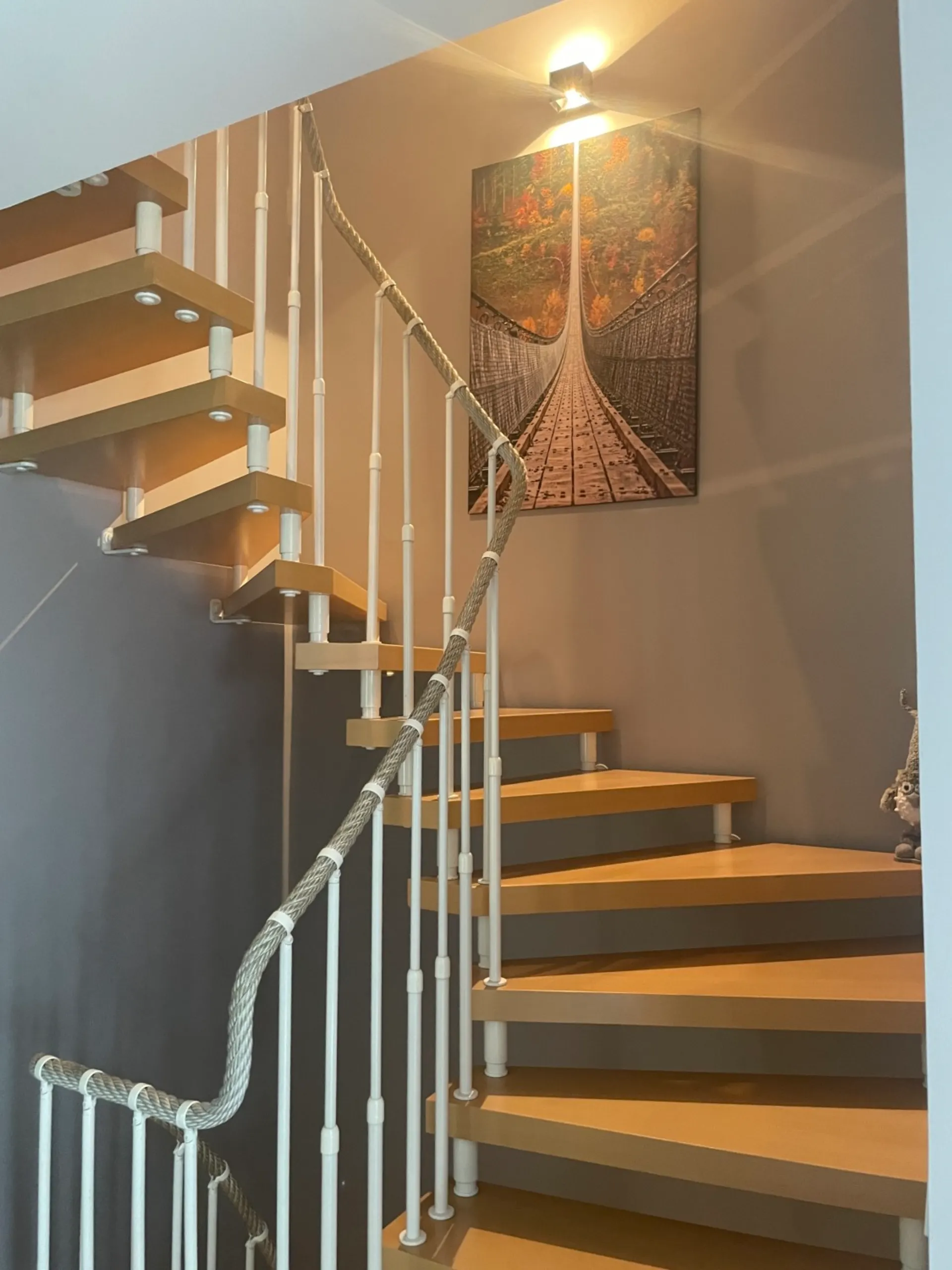 Innenansicht einer Holztreppe mit weißem Geländer und Wandbild einer Hängebrücke.