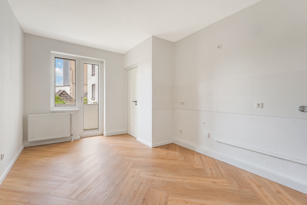 Leerer, heller Raum mit Holzfußboden, Fenster, Tür zum Balkon und weiß gestrichenen Wänden.