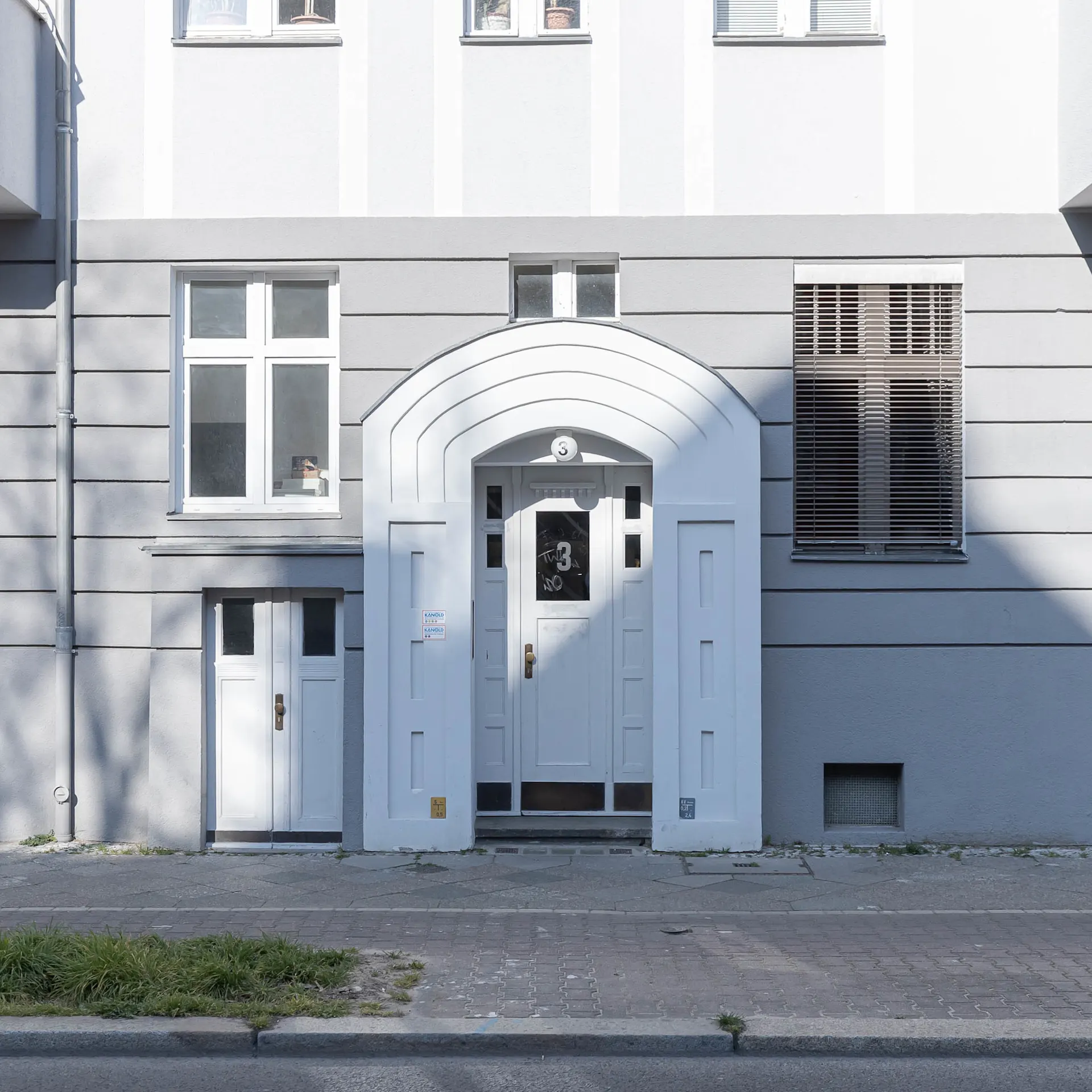 Eingangstür eines Wohngebäudes mit mehreren Fenstern und grauer Fassade.
