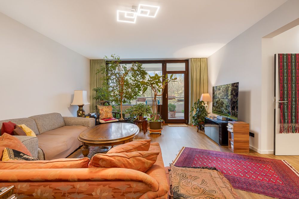 Helles Wohnzimmer mit Sofas, großem Holztisch, vielen Pflanzen und Flachbildfernseher vor Balkon.