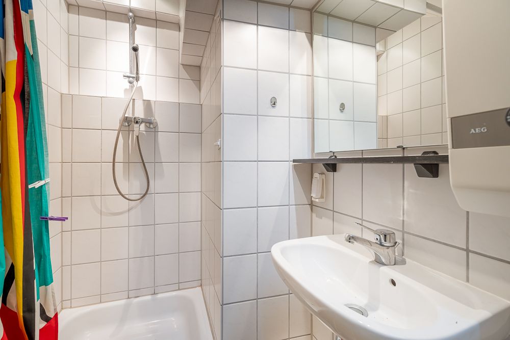 Gefliestes Badezimmer mit Dusche, Waschbecken, Spiegel und buntem Duschvorhang.