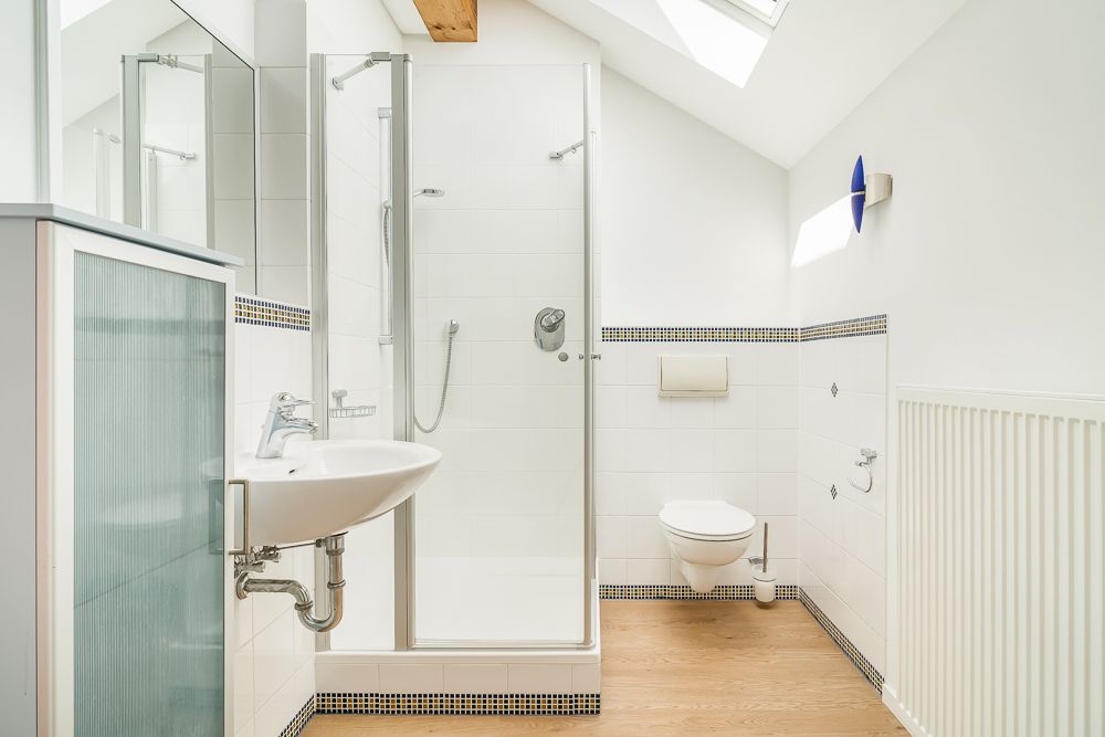Helles Badezimmer mit Dusche, Waschbecken und Wandtoilette, Dachfenster und Holzfußboden.