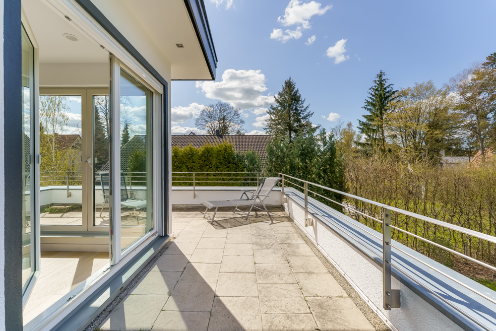 Große Terrasse mit Fliesenboden, zwei Liegestühlen und Blick auf Bäume, Glas-Schiebetüren offen.