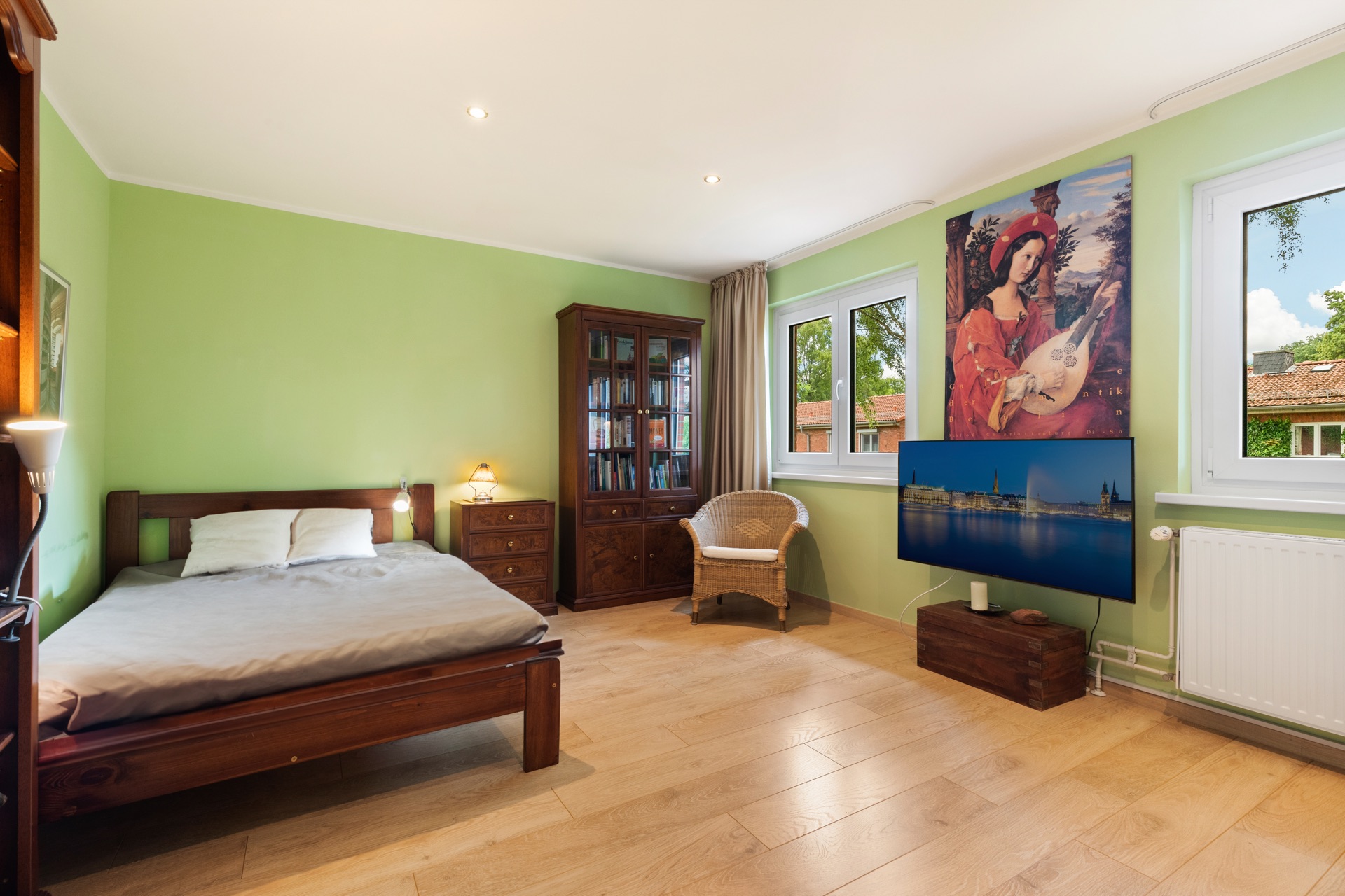 Helles Schlafzimmer mit Holzbett, Sessel, Bücherregal und Fernseher an hellgrüner Wand.