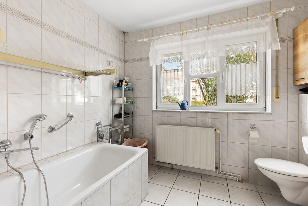 Helles Bad mit Badewanne, Fenster mit Gardinen und Regal neben der Wanne.