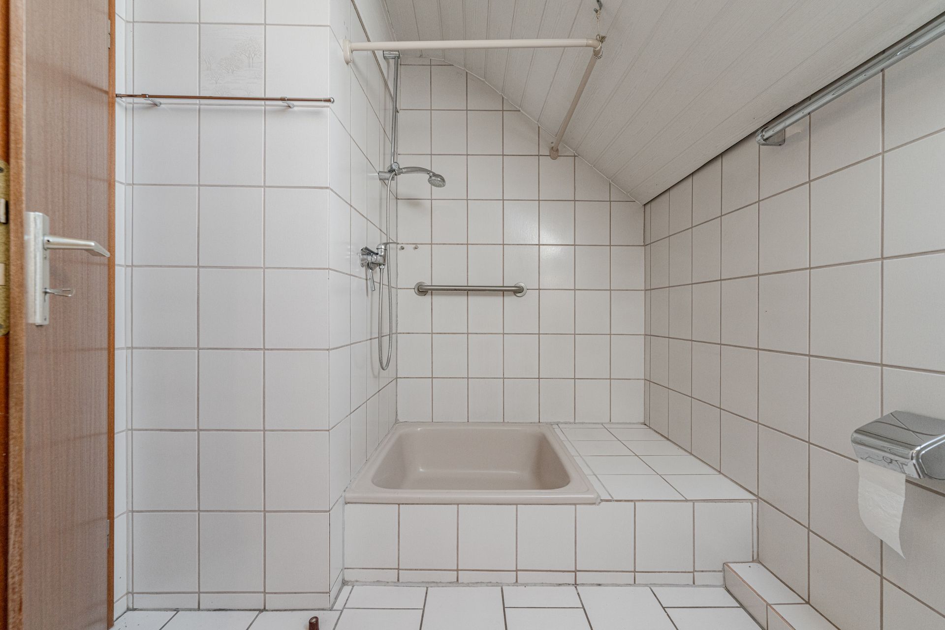 Gefliestes Badezimmer mit Sitzbadewanne, Haltegriff und Duschbrause unter Dachschräge.