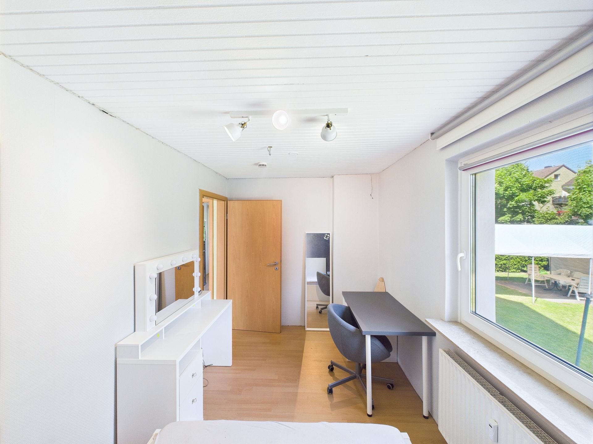 Helles Zimmer mit Schreibtisch, Stuhl, Kommode, Spiegel und großem Fenster zum Garten.