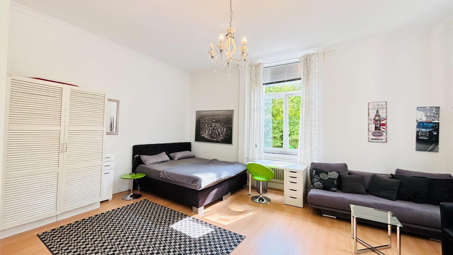 Helles Schlafzimmer mit Doppelbett, Sofa, Schrank, Teppich, Schreibtisch und großem Fenster.