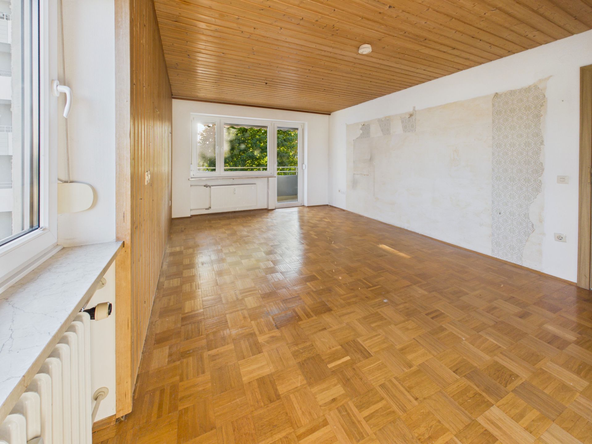 Leerer Raum mit Parkettboden, Holzdecke und großem Fenster zu einem Balkon.