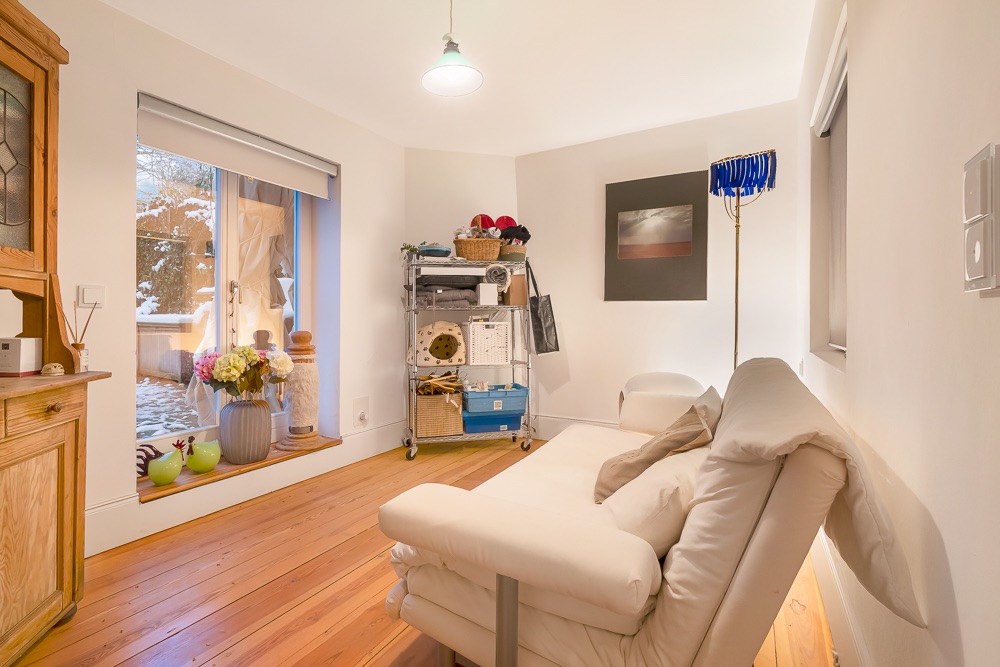 Helles Zimmer mit Sofa, Holzfußboden, Regal, Stehlampe und Fenster mit Blick nach draußen.