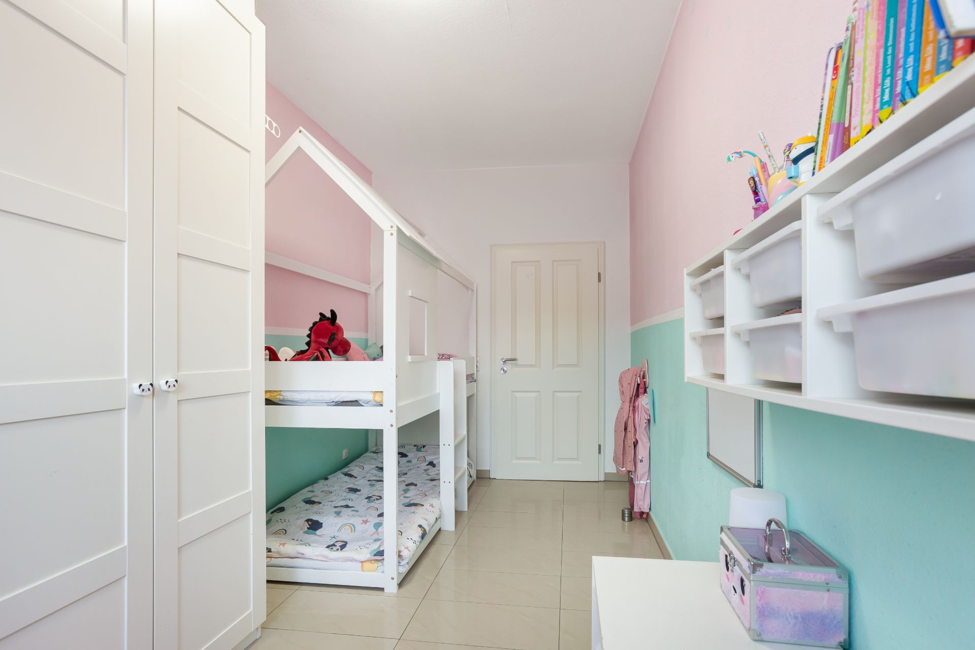 Kinderzimmer mit Etagenbett, Kleiderschrank, Regal mit Büchern und Spielfächern.