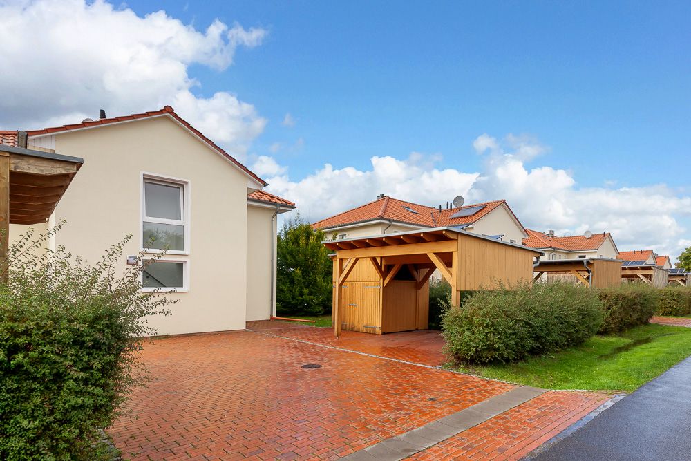 Haus mit rotem Dach, Carport und gepflasterter Einfahrt, umgeben von Büschen.