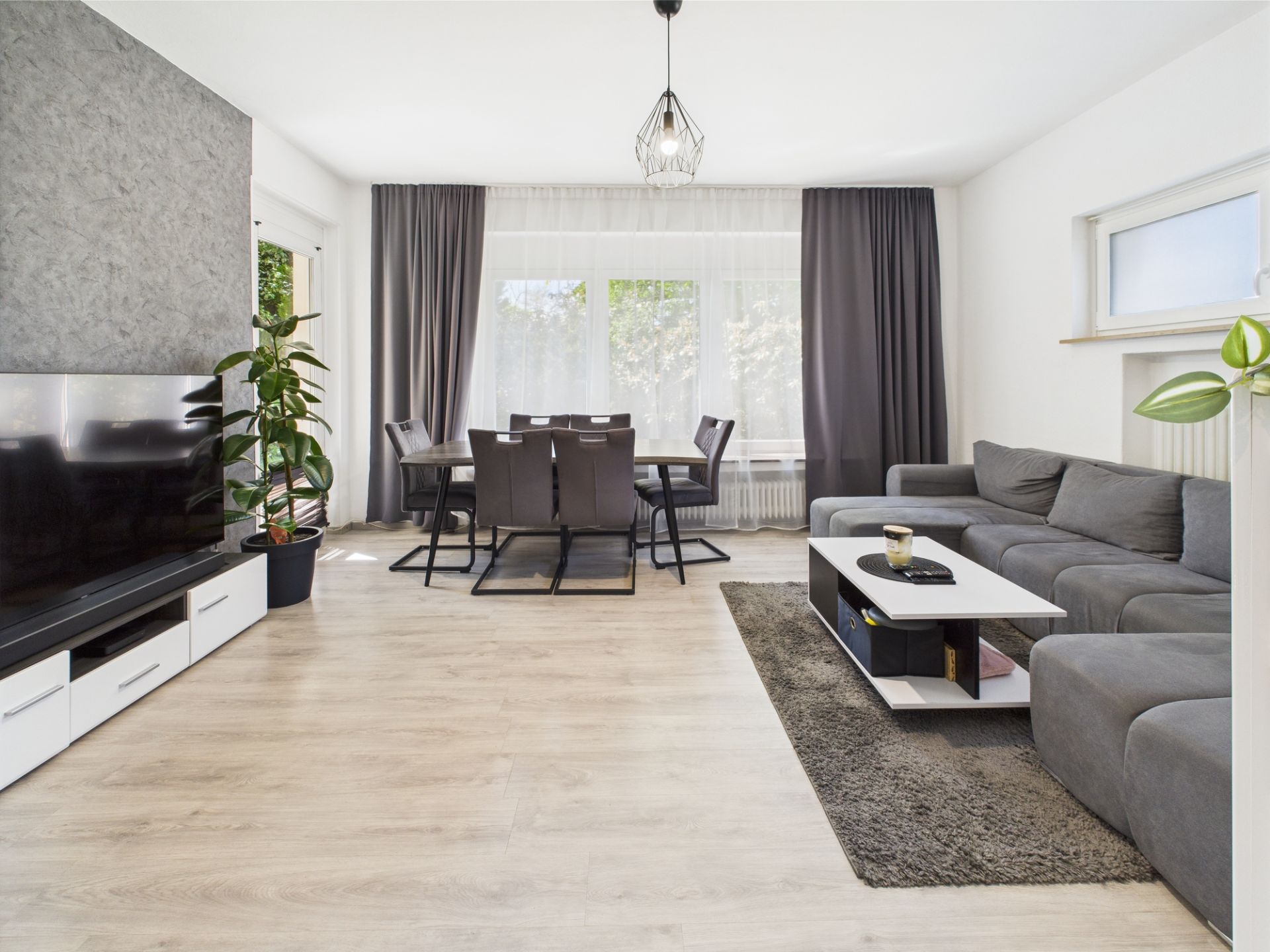 Modernes Wohnzimmer mit grauem Sofa, Fernseher, Esstisch und großen Fenstern mit Vorhängen.