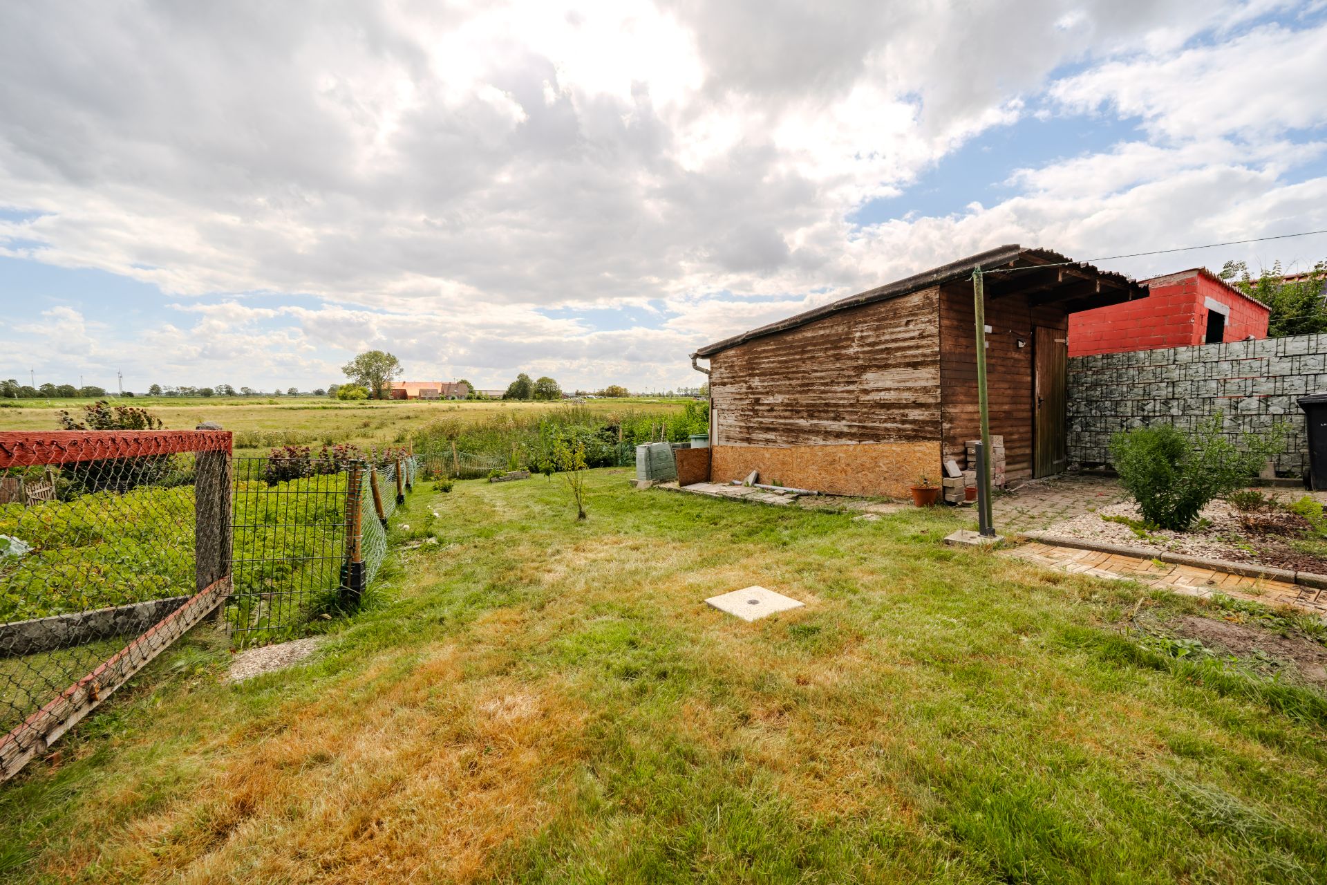 Garten mit Rasenfläche, Holzhütte und Zaun, im Hintergrund Felder und Himmel.