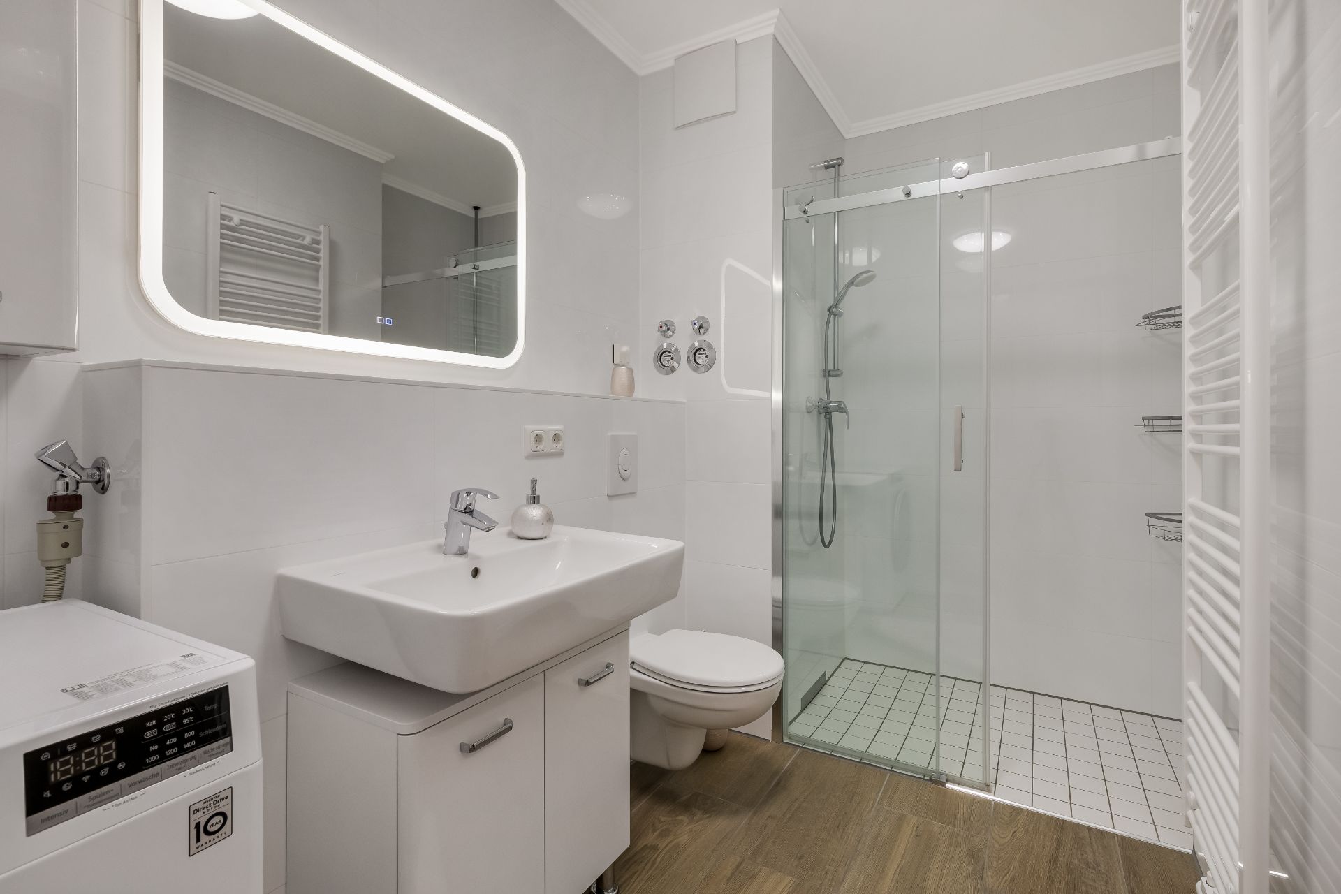 Modernes Badezimmer mit beleuchtetem Spiegel, Waschbecken, Dusche und Waschmaschine.