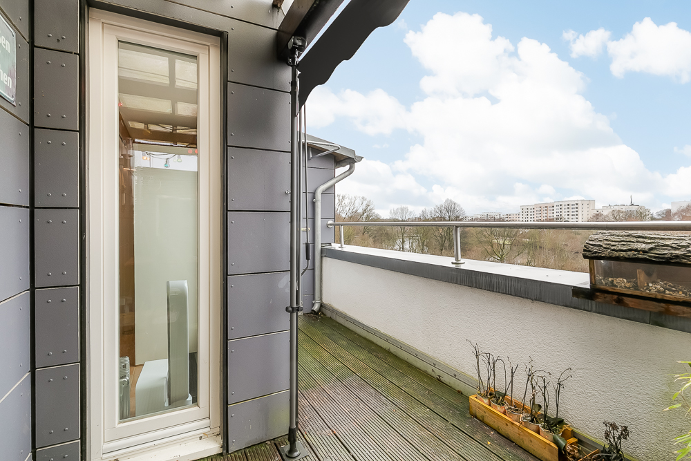 Balkon mit Holzboden, Pflanzkasten und Blick auf Bäume und Wohngebäude im Hintergrund.