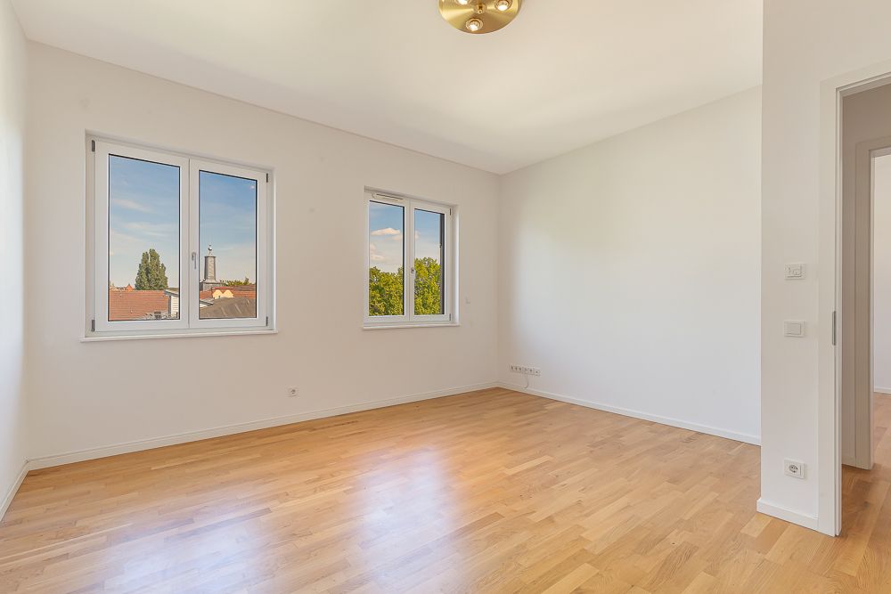 Helles, leeres Zimmer mit Holzboden, zwei Fenstern und weiß gestrichenen Wänden.