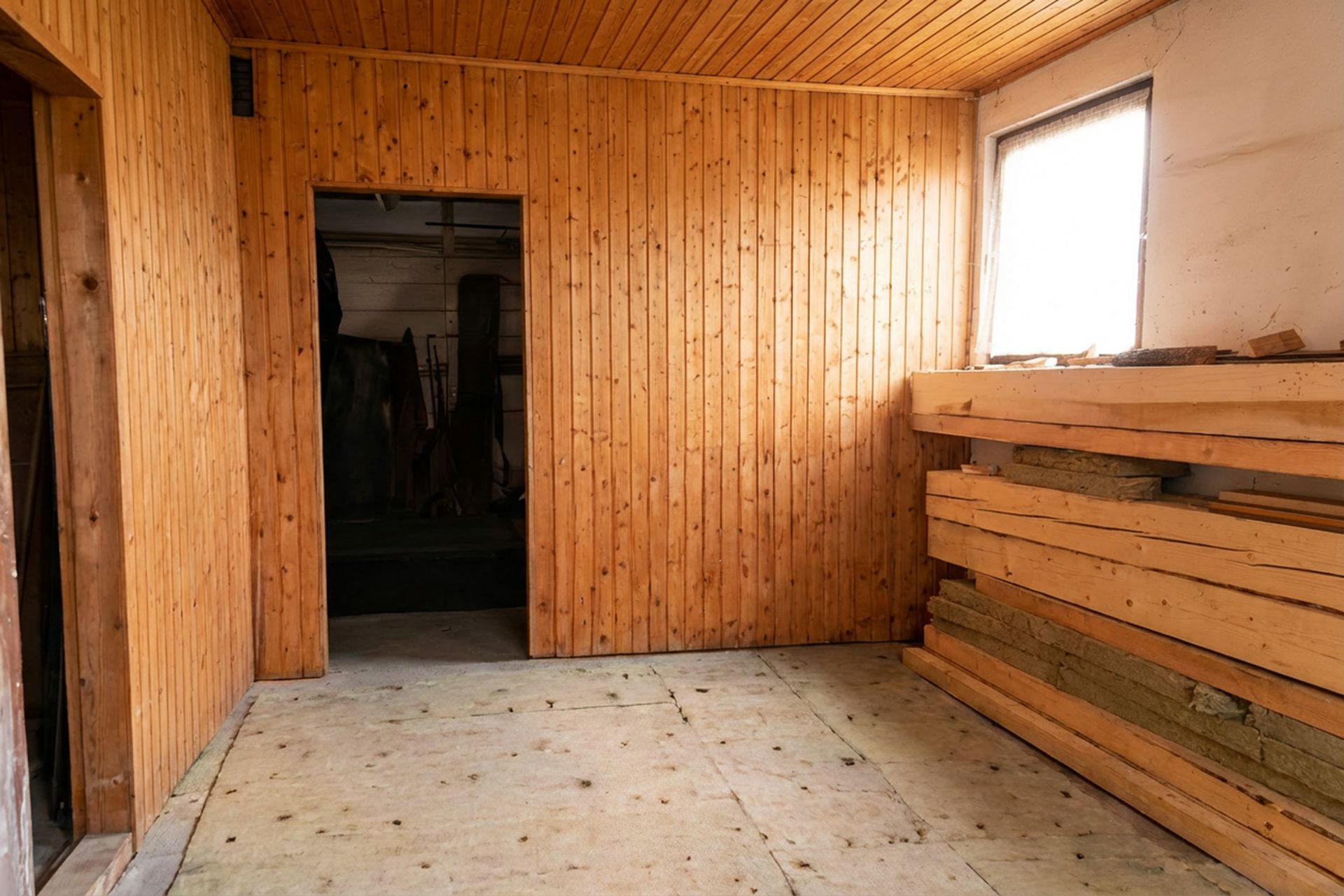 Raum mit Holzverkleidung, Stapel Bretter an der rechten Wand, Fenster rechts.