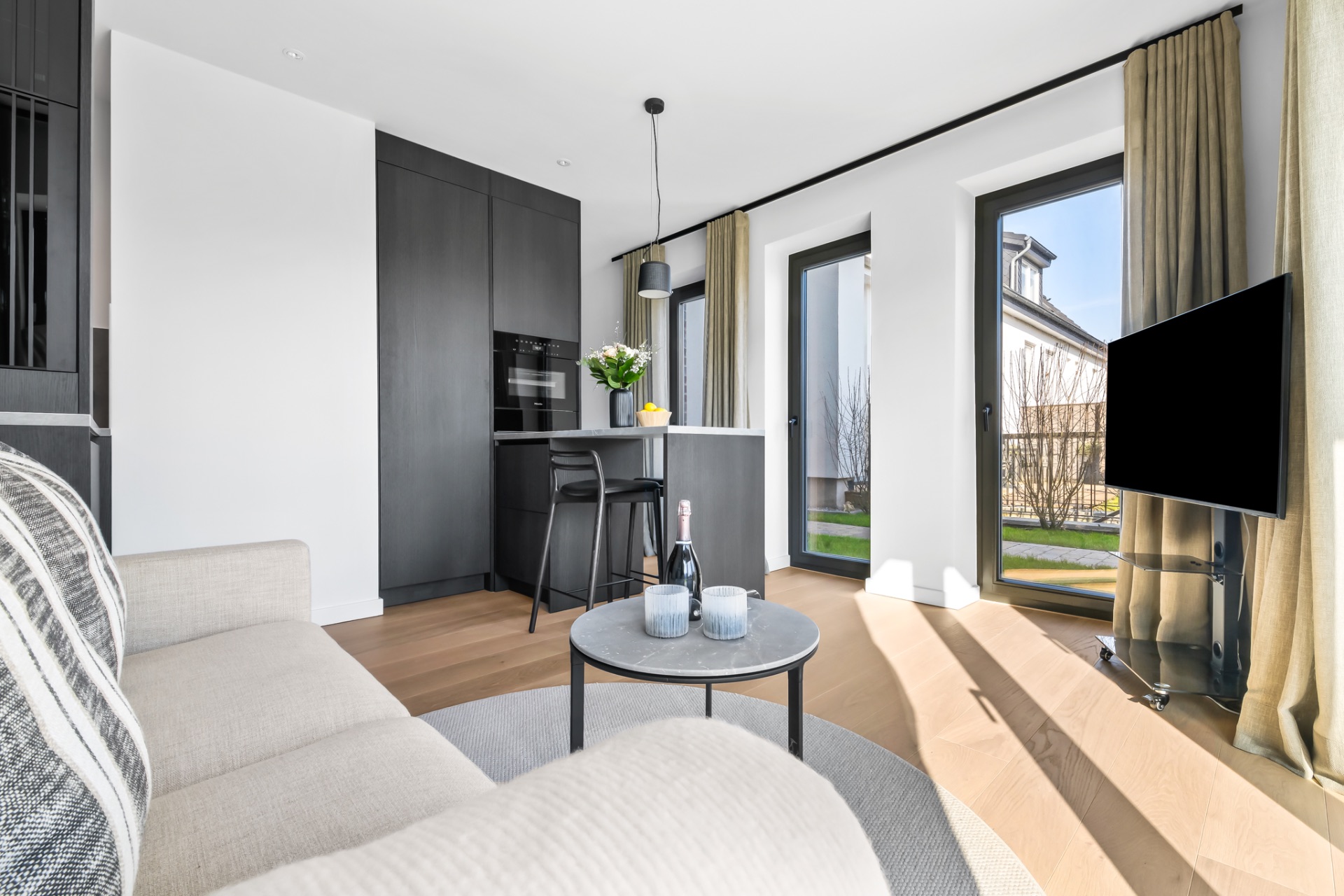 Helles Wohnzimmer mit Sofa, kleinem Tisch, moderner Küche, bodentiefen Fenstern und Fernseher.