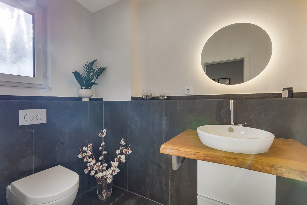 Modernes Badezimmer mit rundem Spiegel, Waschbecken und Toilette, Dekorationen auf dunklen Fliesen.