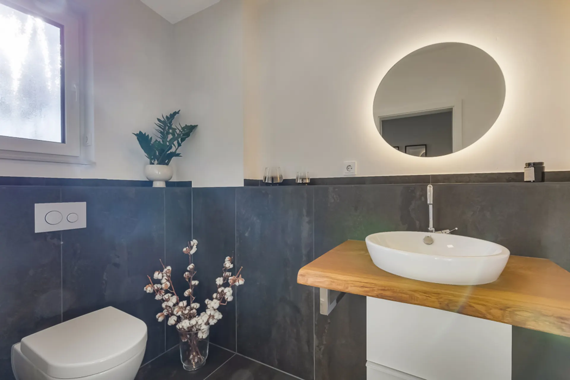 Modernes Badezimmer mit rundem Spiegel, Waschbecken und Toilette, Dekorationen auf dunklen Fliesen.