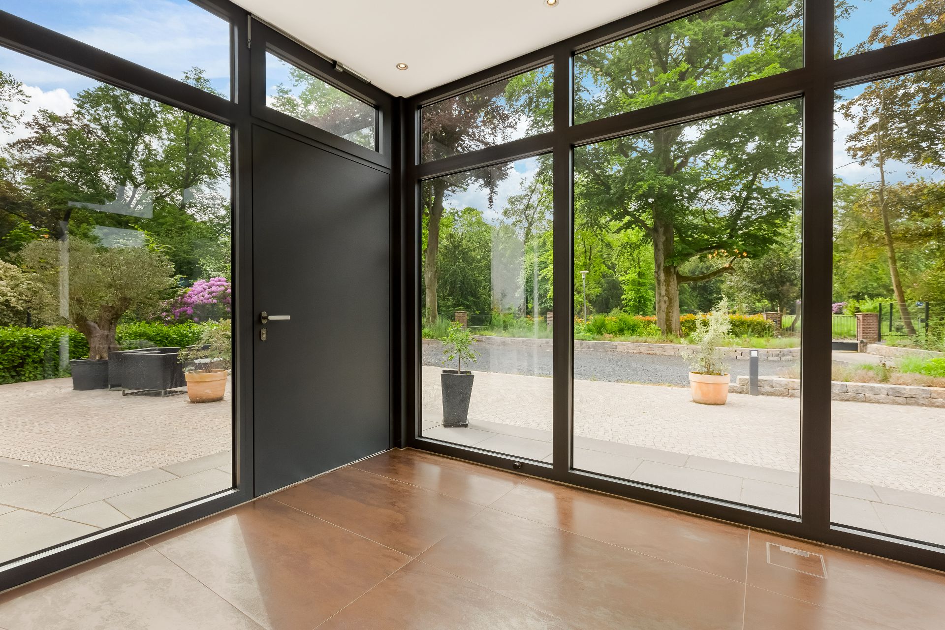 Innenraum mit großen Glasfronten und Blick auf Garten mit Bäumen und Pflanzkübeln.
