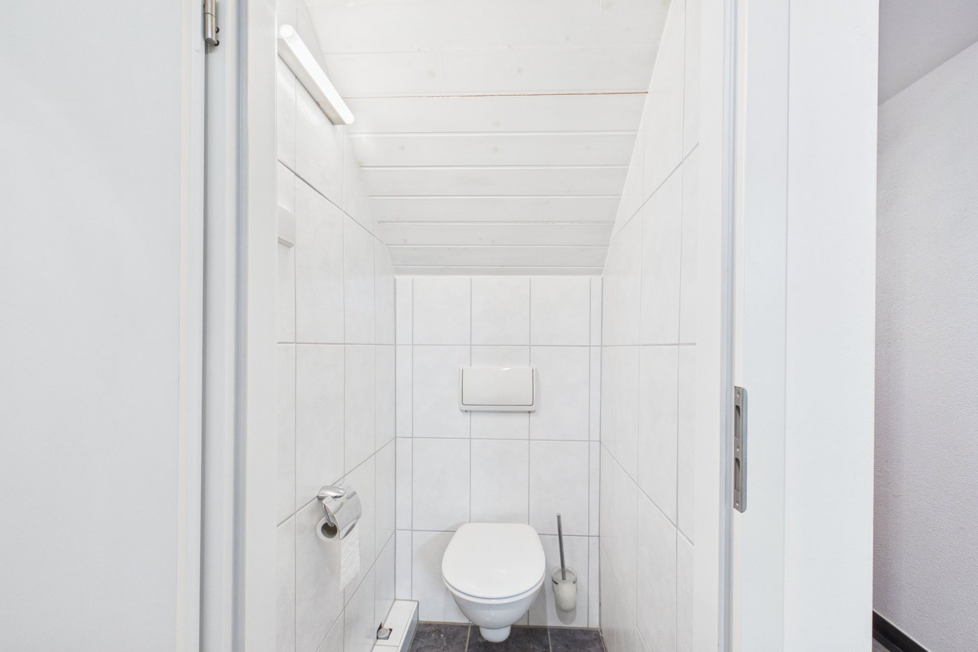 Kleine, weiß geflieste Toilette mit Wand-WC, Toilettenpapierhalter und Bürste unter einer Dachschräge.