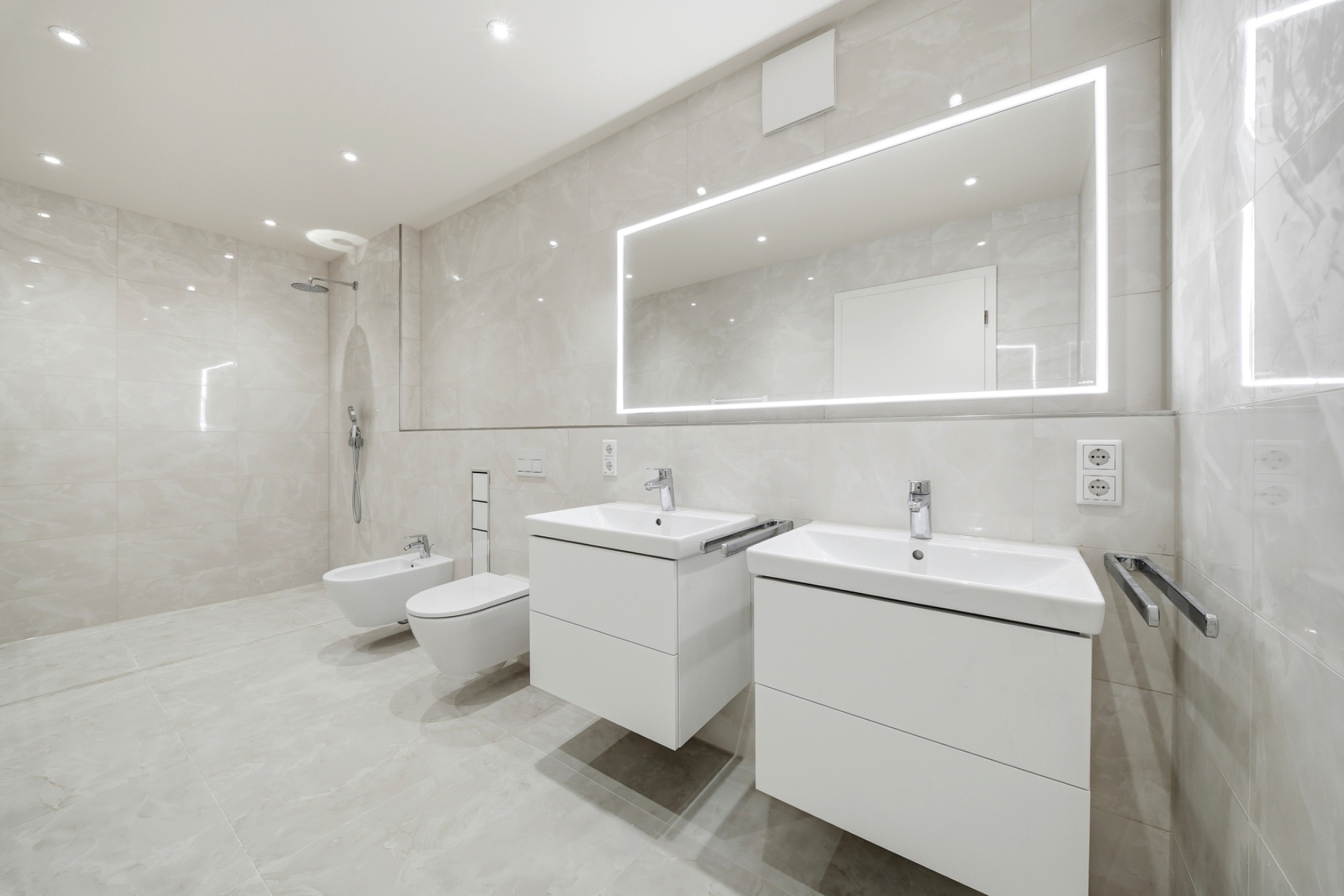 Modernes, helles Badezimmer mit zwei Waschbecken, beleuchtetem Spiegel, WC, Bidet und offener Dusche.