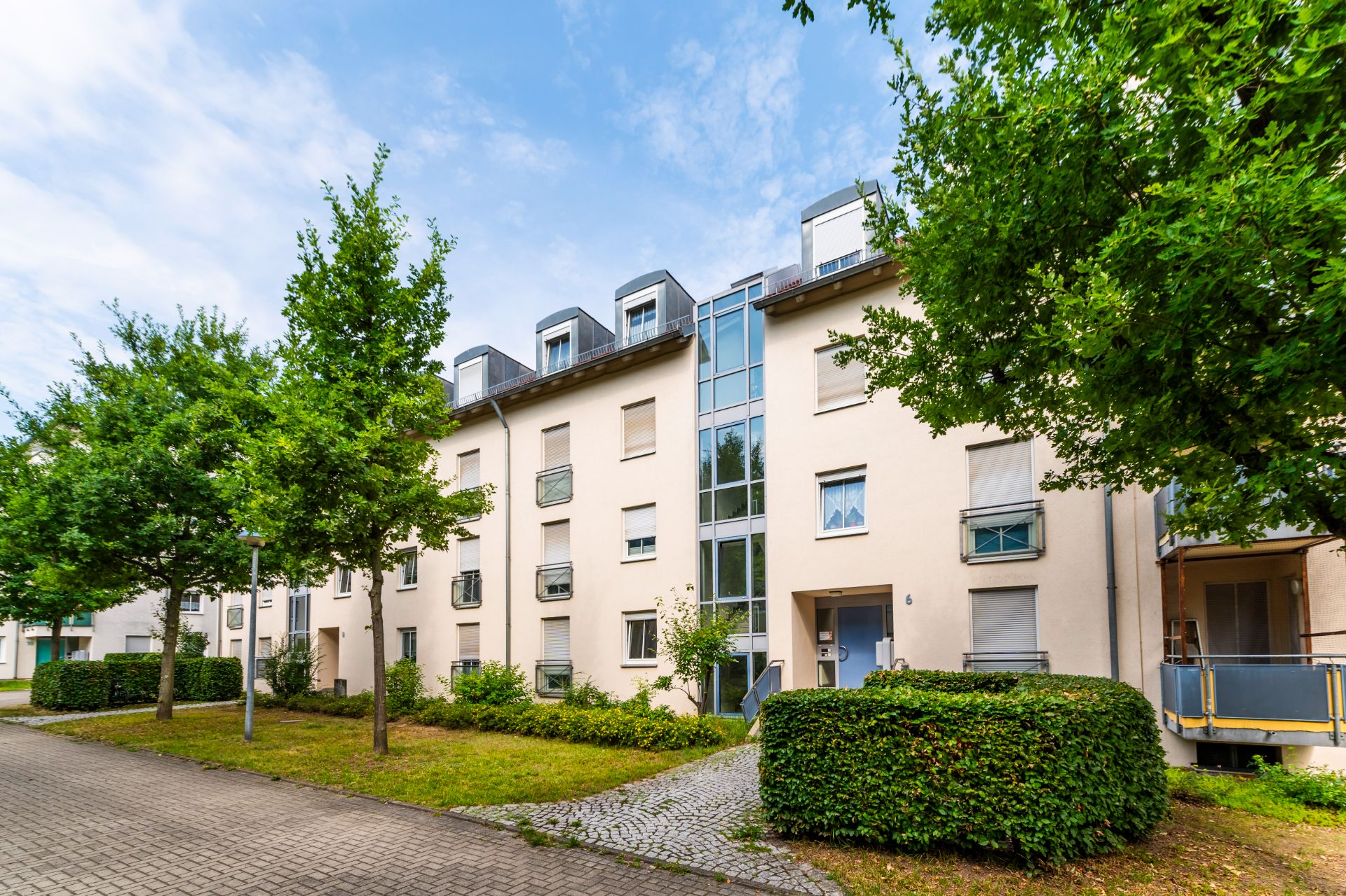 Mehrstöckiges Wohngebäude mit Bäumen, Gehweg und grüner Bepflanzung davor.