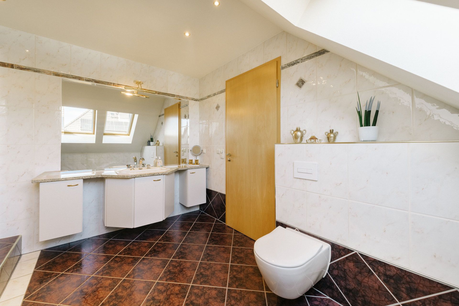 Modernes Badezimmer mit WC, Doppelwaschbecken, Spiegel und Pflanzen auf braunem Fliesenboden.