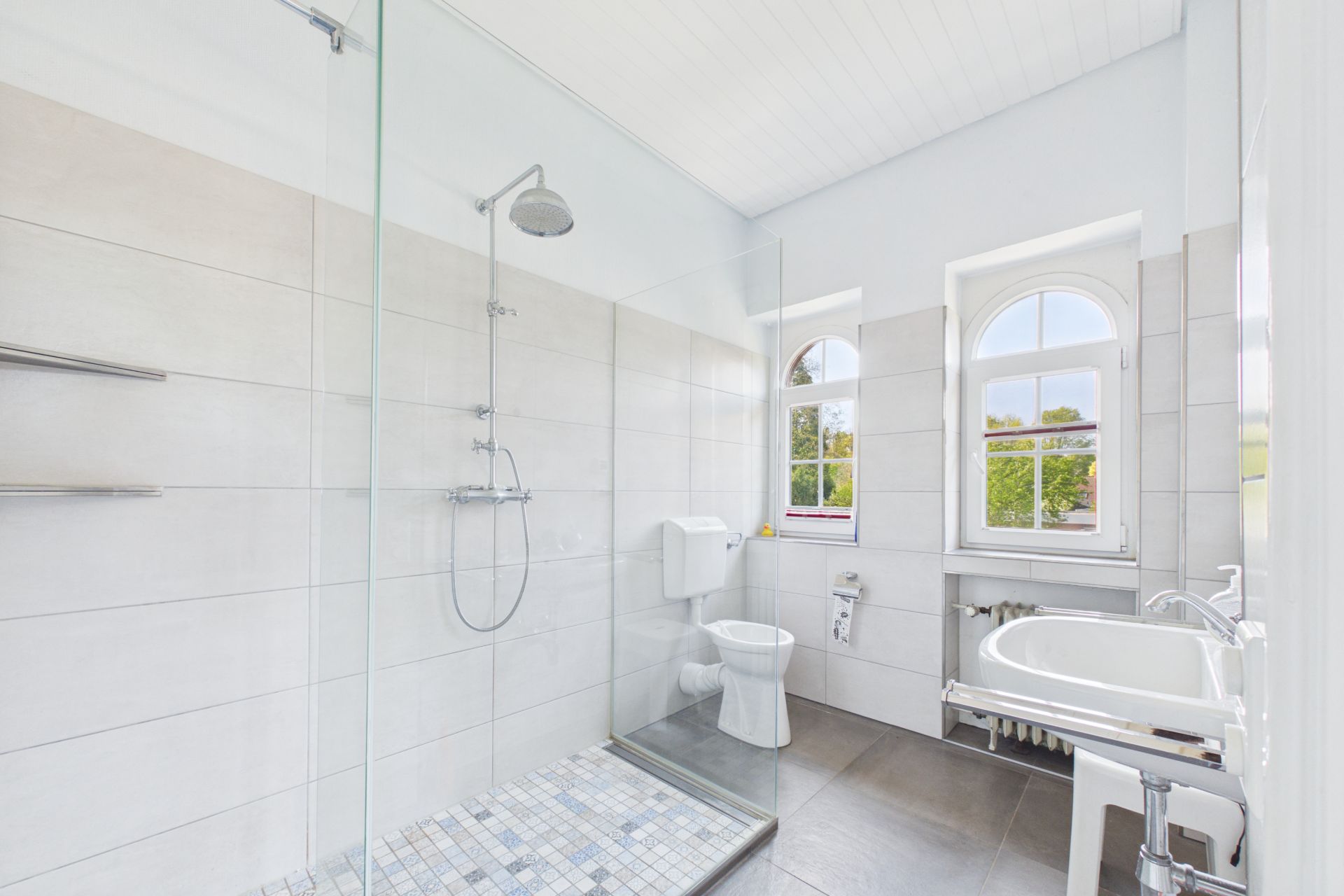 Helles Badezimmer mit bodengleicher Dusche, WC, Waschbecken und zwei Fenstern.