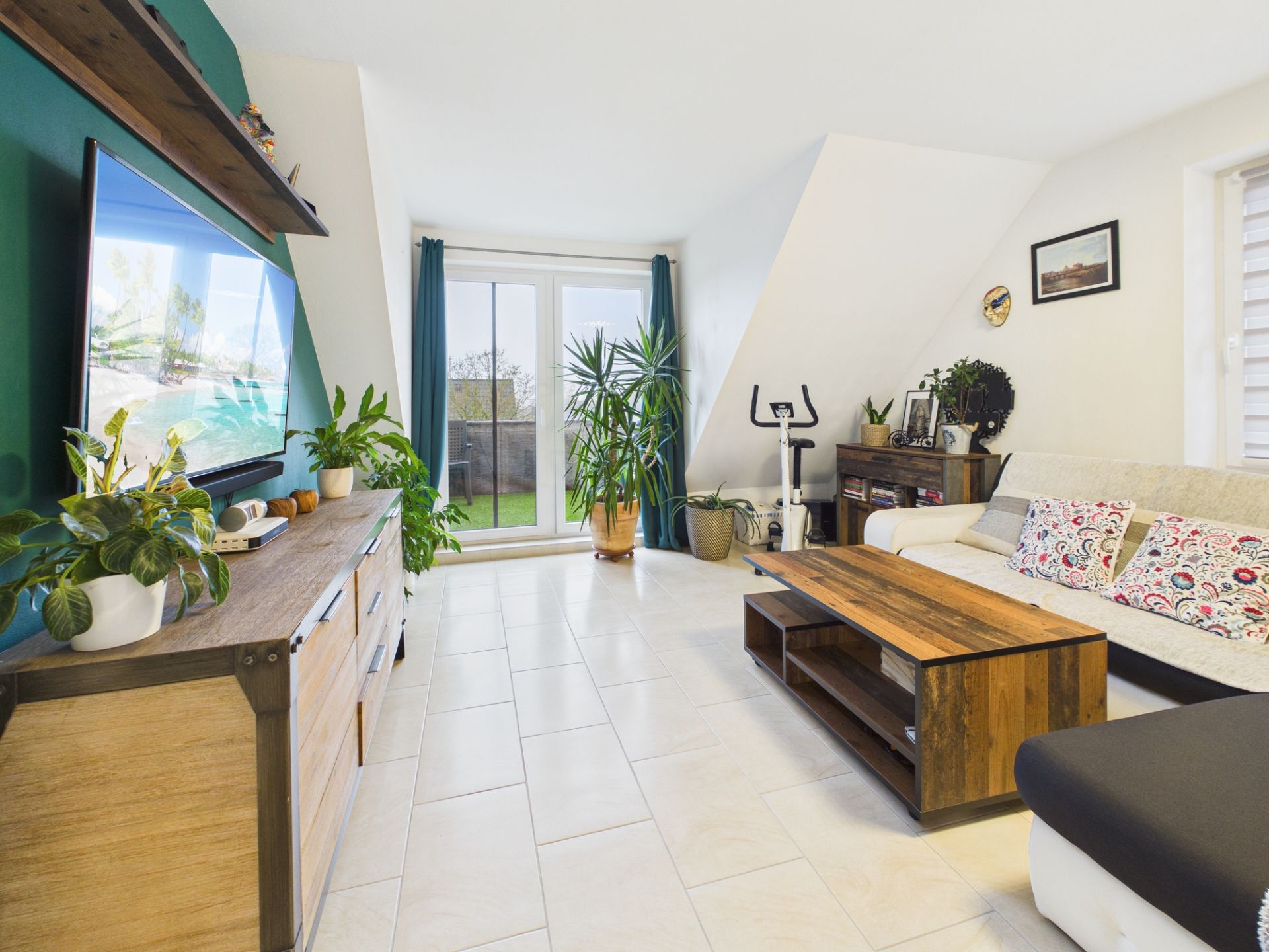 Helles Wohnzimmer mit Sofa, Couchtisch, Fernseher, Pflanzen und großem Fenster mit Vorhängen.