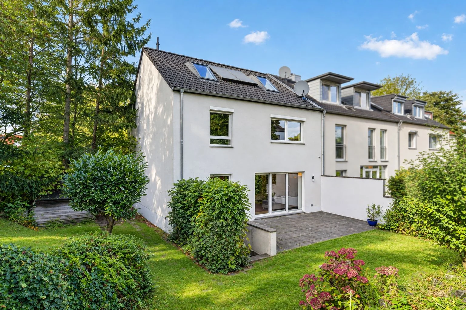 Helles Wohnhaus mit Terrasse und Garten, umgeben von Büschen und Bäumen.