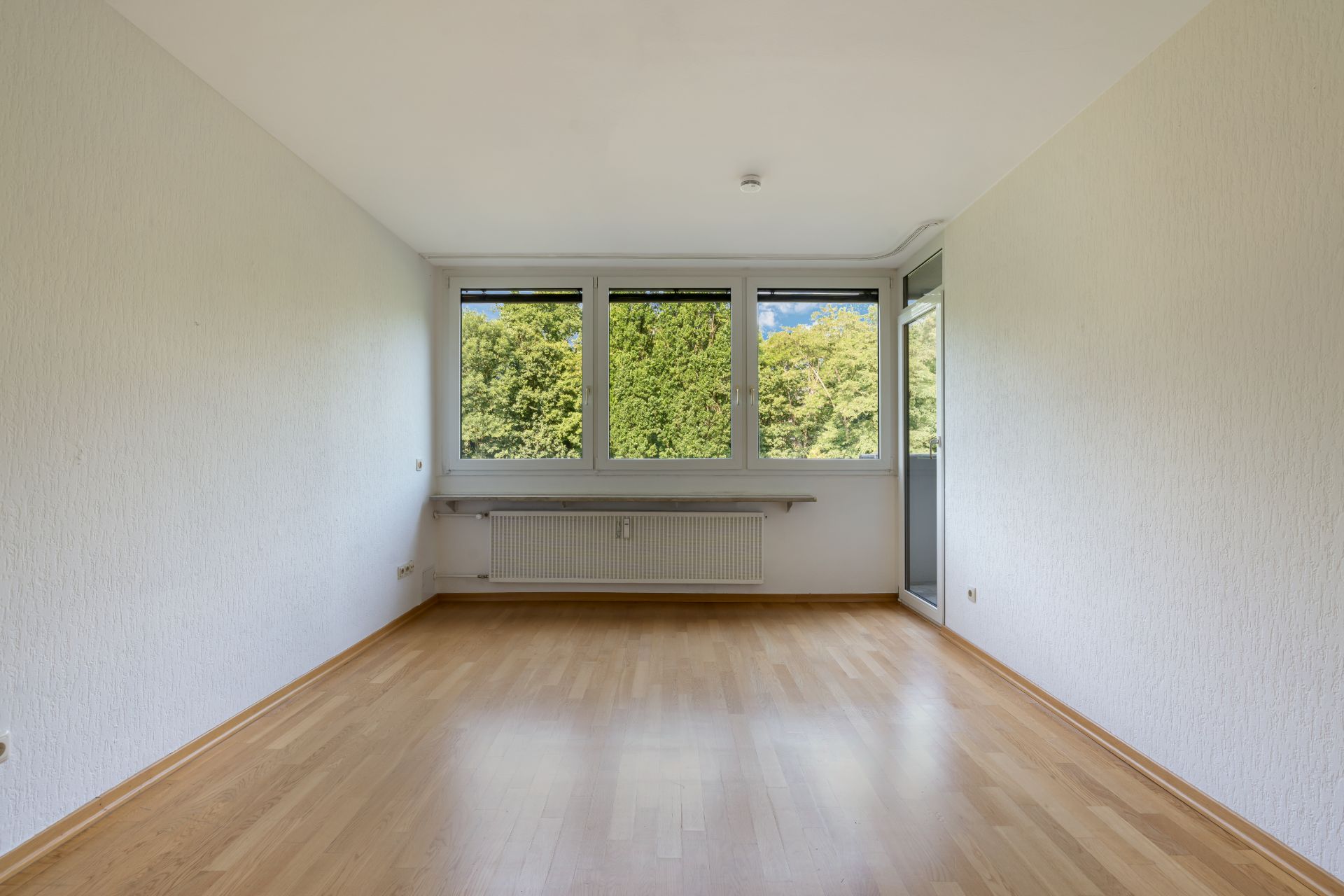 Leerer Raum mit großen Fenstern, Parkettboden und Blick ins Grüne.