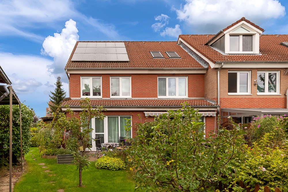 Rotes Reihenhaus mit Solarpanels auf dem Dach und grünem Garten im Vordergrund.
