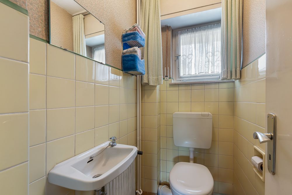 Kleines Badezimmer mit Toilette, Waschbecken und Fenster mit Gardine.