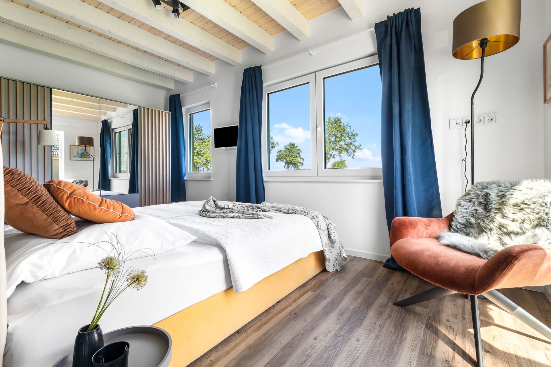 Helles Schlafzimmer mit großem Bett, Sessel, Stehlampe und Fenstern mit blauen Vorhängen.