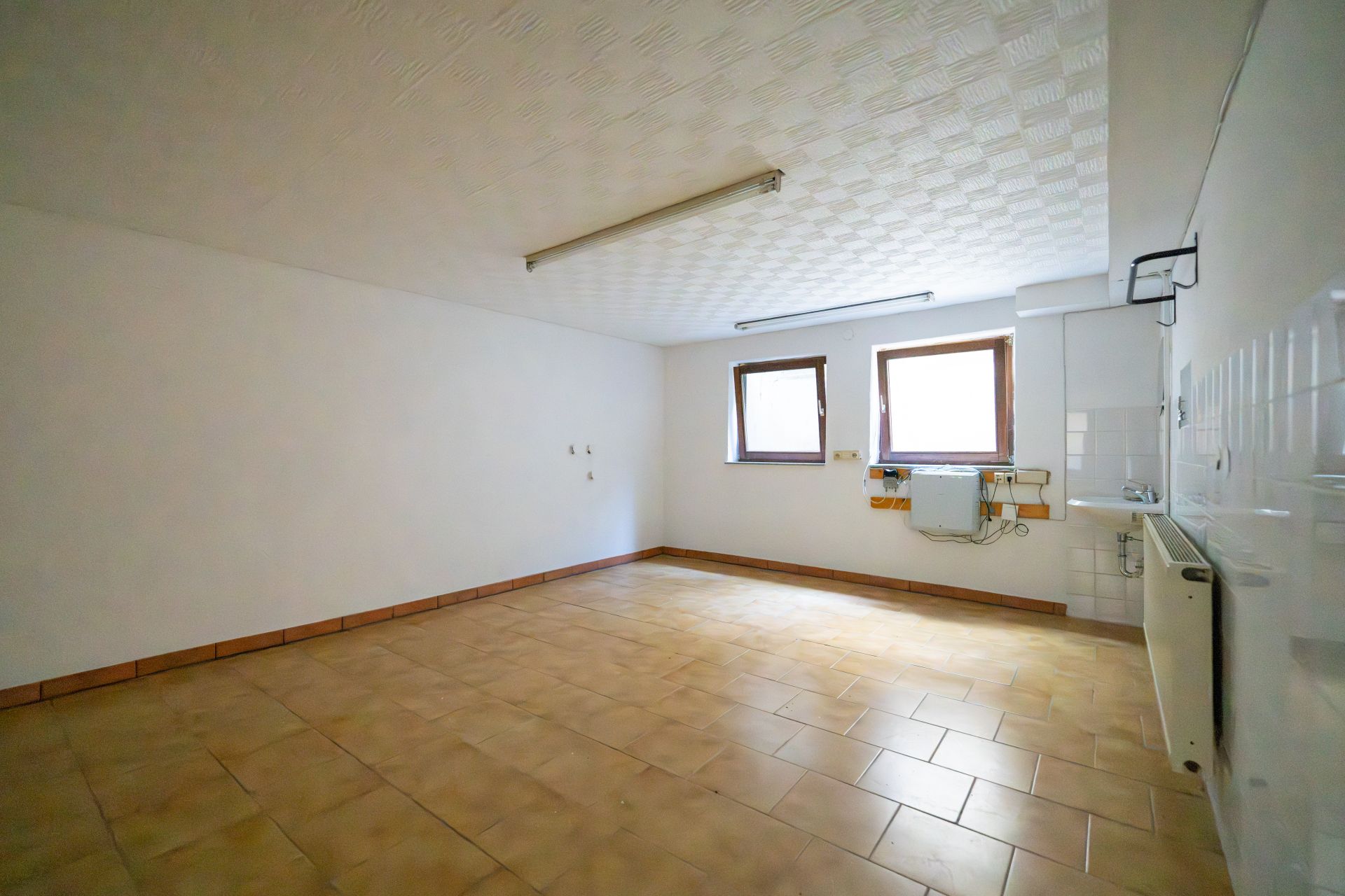 Leerer Raum mit zwei Fenstern, gefliestem Boden und Heizkörper an der Wand.