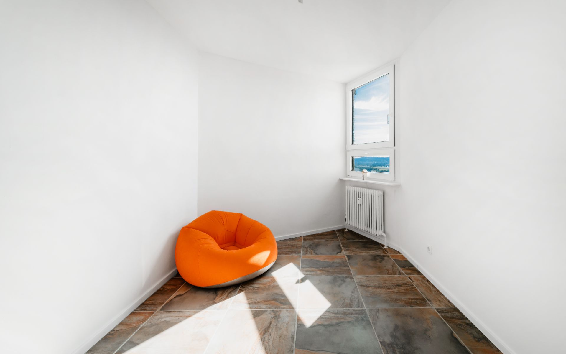 Kleiner, heller Raum mit orangefarbenem Sitzsack, Fliesenboden und Fenster mit Ausblick.