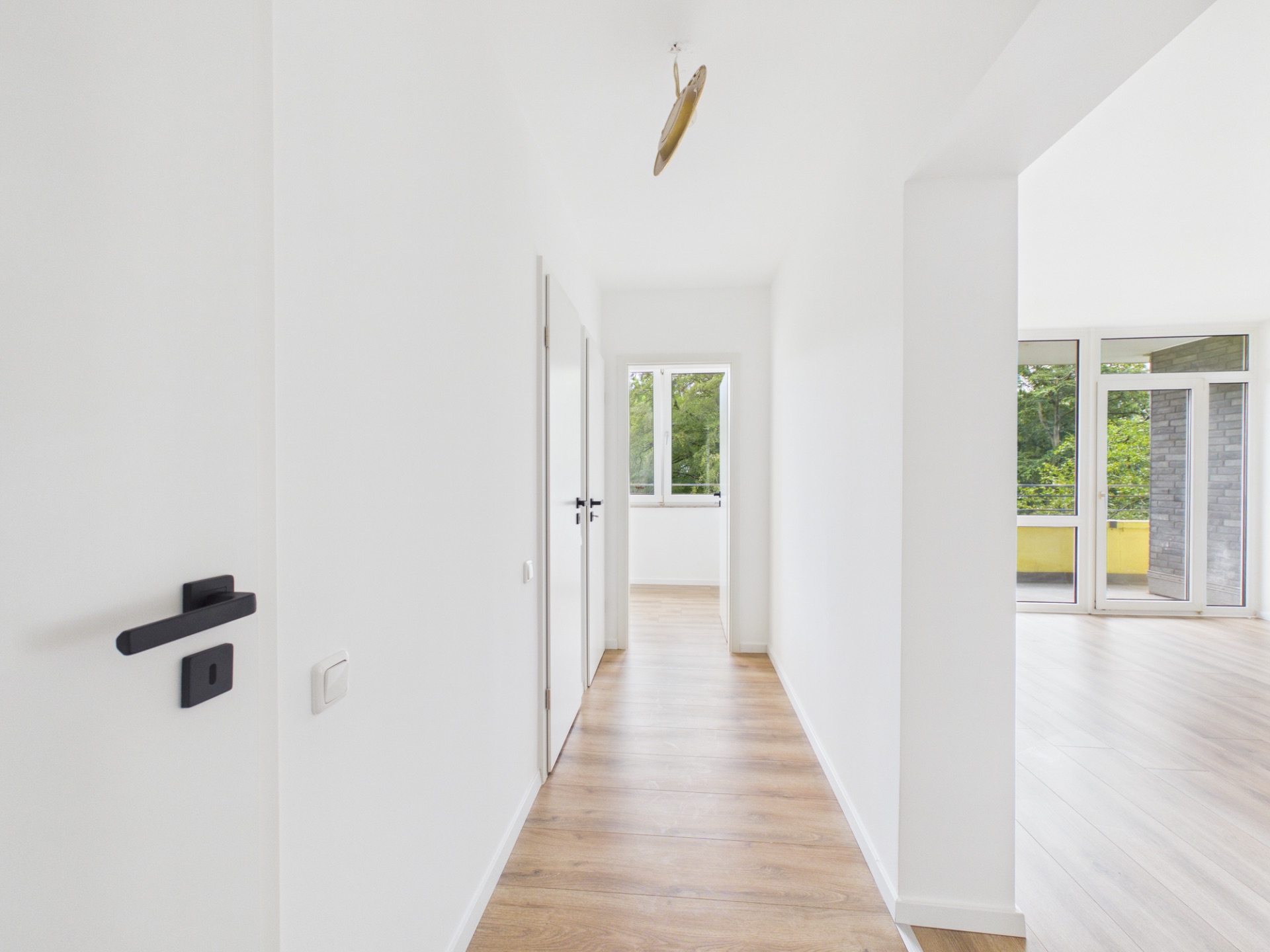 Heller Flur mit Holzboden, weißen Wänden und Türen, Fenster am Ende, Zugang zu einem Raum rechts.
