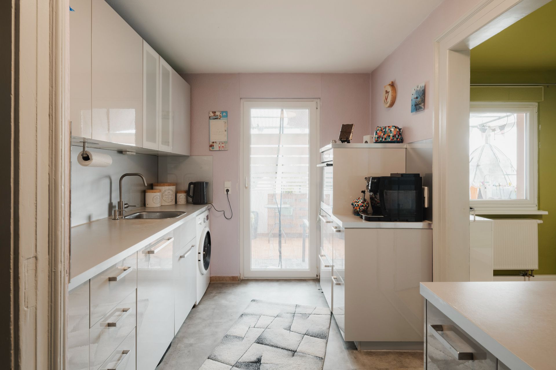 Moderne Küche mit weißen Schränken, Kaffeemaschine und Zugang zur Terrasse durch Glastür.