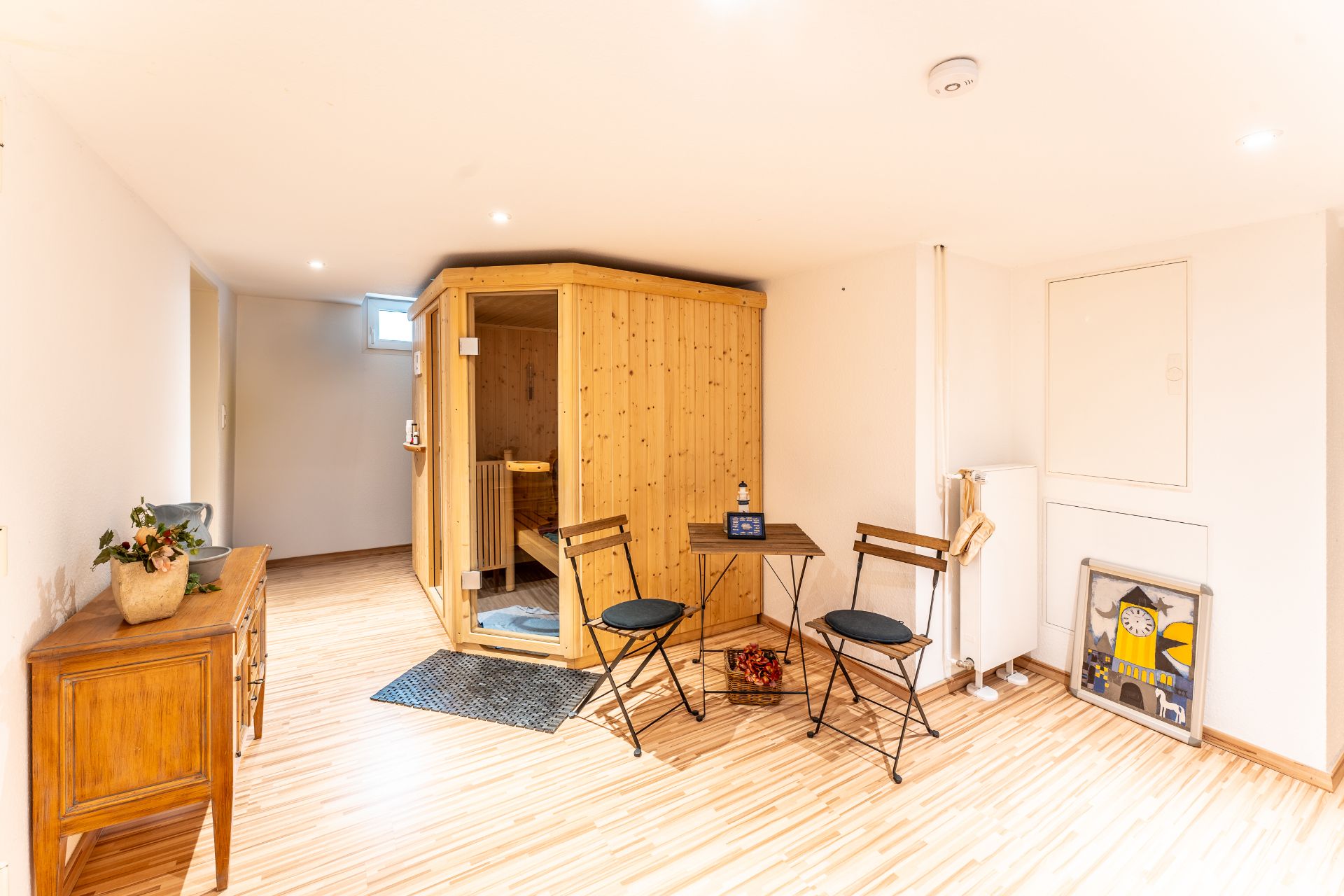 Heller Raum mit Holzsauna, kleinem Tisch, zwei Stühlen und Sideboard auf Holzfußboden.
