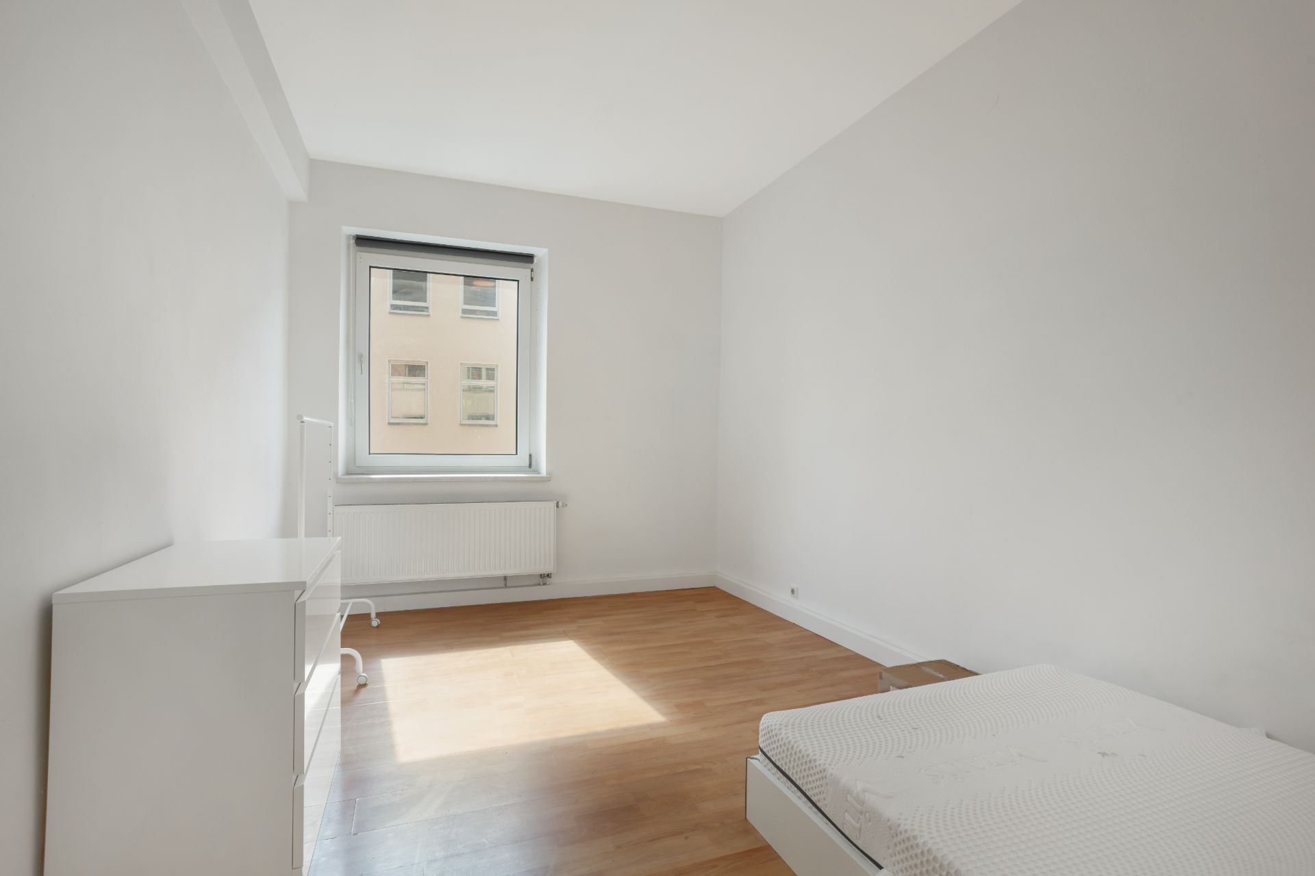Helles Schlafzimmer mit Bett, Kommode und Fenster, Holzboden und weißen Wänden.