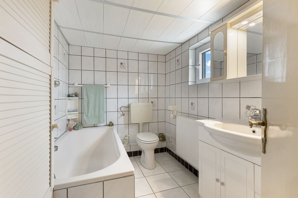 Helles Badezimmer mit Badewanne, Toilette, Waschbecken und Spiegelschrank, gefliest mit weißen Fliesen.