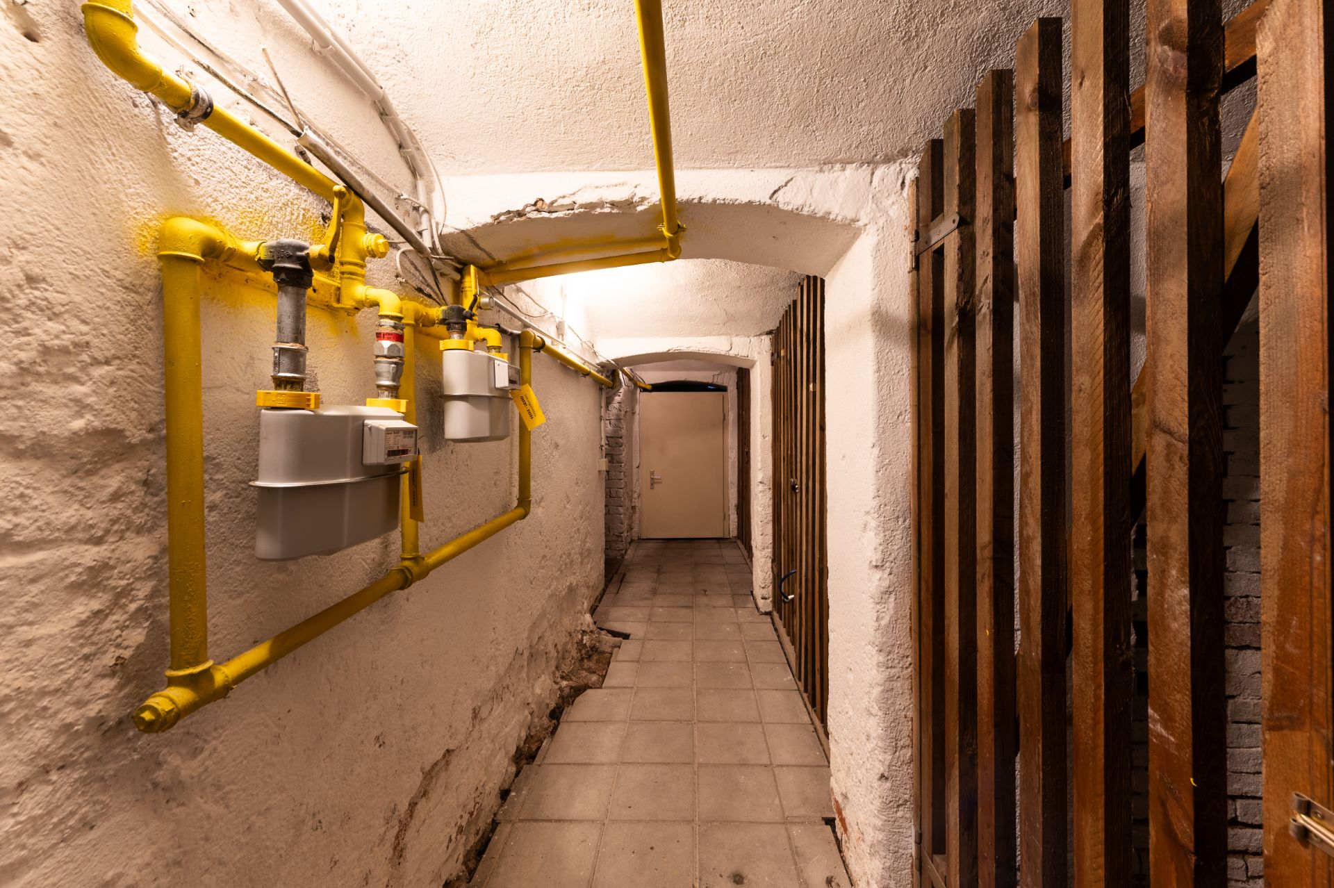 Kellerflur mit gelben Gasleitungen und Holzgitter an der Wand.