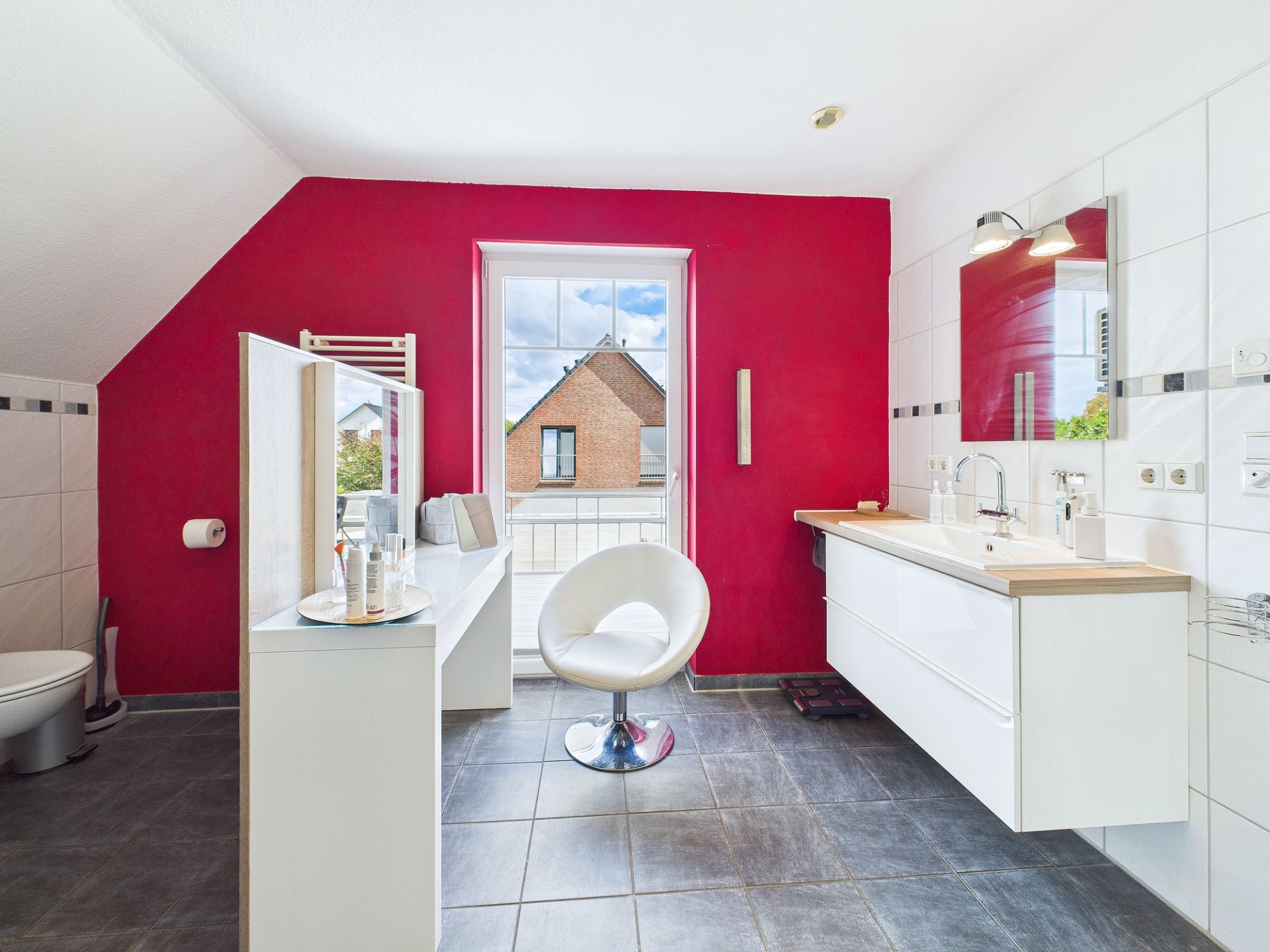 Modernes Badezimmer mit roter Wand, großem Fenster, weißem Waschbecken, Spiegel und drehbarem Stuhl.
